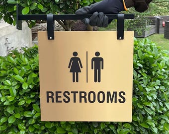Peut inclure: Panneau de toilettes doré avec texte et symboles noirs. Le panneau présente des silhouettes féminines et masculines, avec le mot « RESTROOMS » en dessous. Le panneau est suspendu à un support en métal noir.