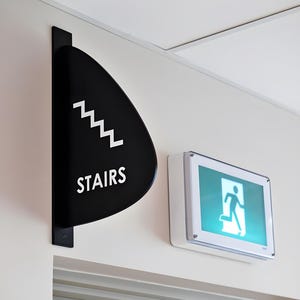 May include: A black and white sign with the word "STAIRS" and a white staircase symbol. The sign is mounted on a white wall next to a green emergency exit sign with a running person symbol.