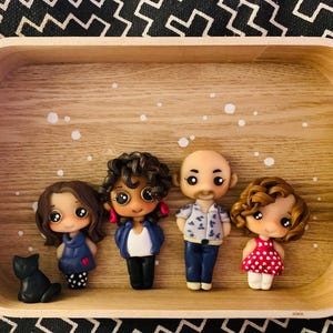 以下が含まれることがあります： 木製の表面に並んだ、手作りのポリマークレイ製の4体の人の人形。茶色の髪の女の子が青いドレスを着て、茶色の髪の女の子が赤いドレスを着て、茶色の髪の女の子が青いシャツを着て、茶色の髪の男性が青いシャツを着ています。