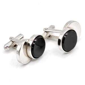 May include: A pair of silver-toned cufflinks featuring a circular design with a smooth, polished black inlay at the center. Each cufflink has a hinged toggle mechanism on the back, allowing for secure fastening through shirt buttonholes.
