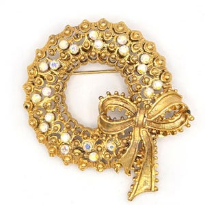 May include: A gold-toned wreath brooch with a bow. The wreath is adorned with clear, iridescent rhinestones. The brooch has a pin closure on the back and is set against a white background.