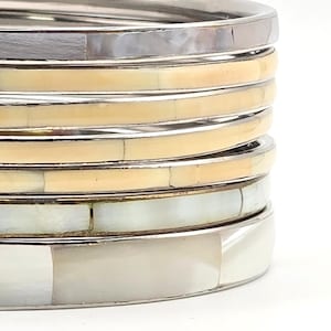 May include: A stack of six circular bangles featuring alternating bands of polished silver-toned metal and inlaid segments of cream, beige, and iridescent white shell. The bracelets are arranged vertically, showcasing a smooth, segmented surface.