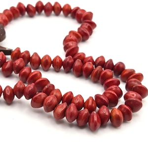 May include: A necklace composed of numerous small, smooth, red, seed-like beads with a pointed, oval shape. The beads are strung together in a continuous loop, displaying a uniform deep red color and a polished texture throughout the entire strand.