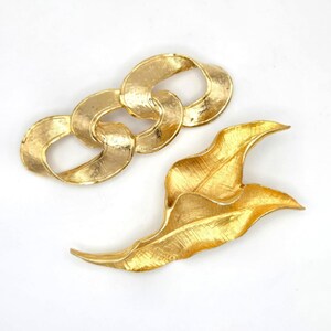 May include: Two gold-toned vintage brooches. One brooch is a chain link design, and the other is a leaf design. Both brooches have a textured surface and are set against a white background.