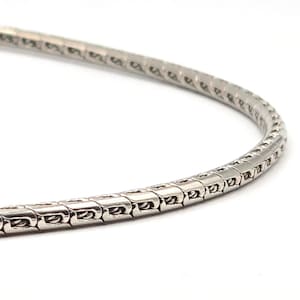 May include: A silver-colored necklace with a snake chain design. The chain is made of small, interconnected rectangular links, creating a flexible and smooth texture. The necklace is curved, suggesting it is being displayed or worn.