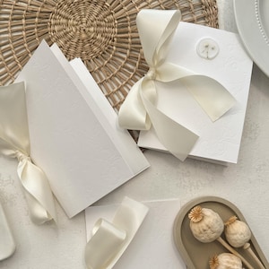 May include: Three white cards with embossed designs and tied with ivory satin ribbon bows. The cards are on a white surface with a woven wicker mat and a small dish with a gold number "3" on it.