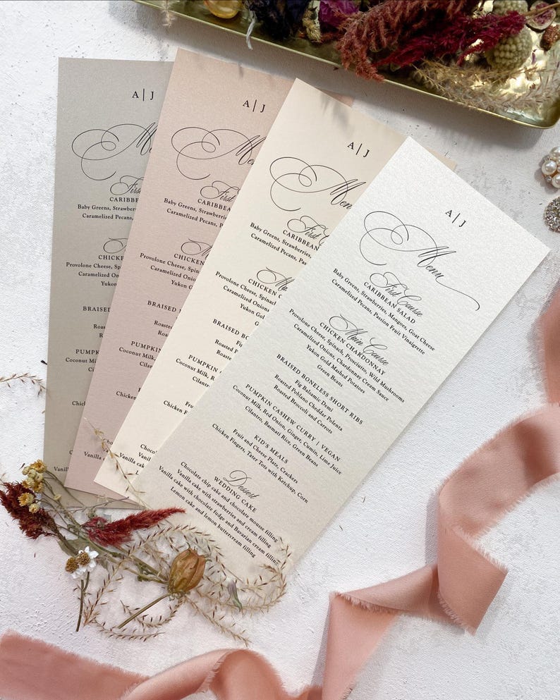 May include: Four beige menu cards with black calligraphy text. The text includes the words 'First Course', 'Caribbean Salad', 'Chicken Chardonnay', 'Braised Boneless Short Ribs', 'Pumpkin Cashew Curry', 'Kids Meals', 'Wedding Cake', and 'Dessert'. The menu cards are stacked on top of each other and are surrounded by pink silk ribbon and dried flowers.