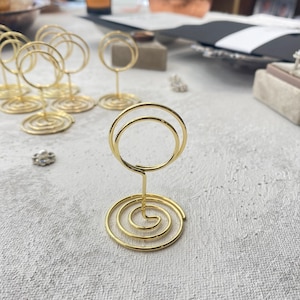 May include: Gold-colored wire place card holders with a spiral base and a double-circle top. Several holders are arranged on a textured, light-colored surface, suggesting a wedding or event setting. A few small decorative items are also visible.
