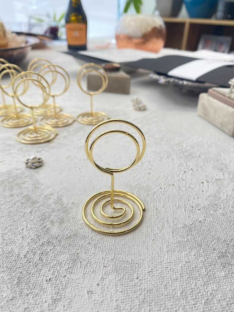 May include: Gold-colored wire place card holders with a spiral base and a double-circle top. Several holders are arranged on a textured, light-colored surface, suggesting a wedding or event setting. A few small decorative items are also visible.