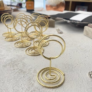 May include: Gold-colored wire place card holders with a spiral base and a circular top. The holders are arranged on a textured, light-colored surface, suggesting a table setting or event decor.