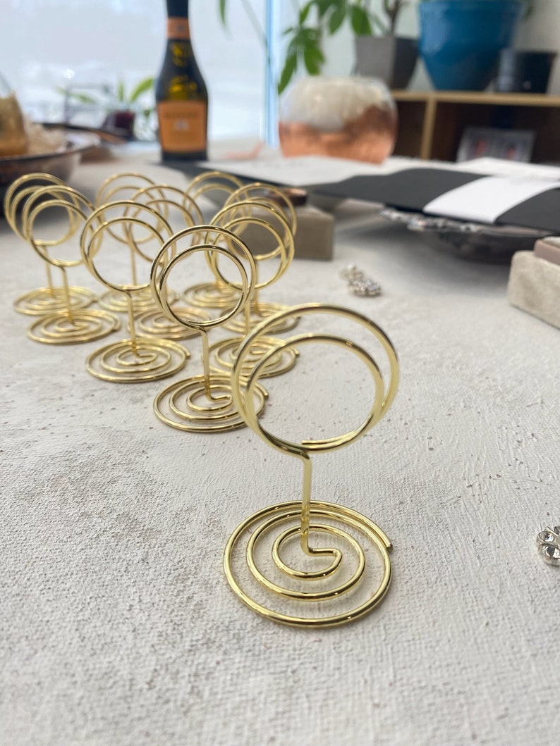 May include: Gold-colored wire place card holders with a spiral base and a circular top. The holders are arranged on a textured, light-colored surface, suggesting a table setting or event decor.