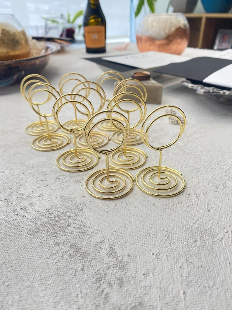 May include: A collection of gold-colored wire place card holders. Each holder features a spiral base and a circular top for holding cards. The holders are arranged on a textured, light-colored surface, suggesting a wedding or event setting.