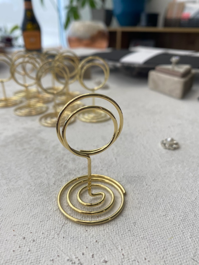 May include: Gold-colored metal place card holders with a spiral base and a double-loop top. The holders are arranged on a textured, light-colored surface, suggesting a wedding or event setting. Other holders are visible in the background.