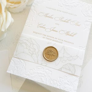 May include: Elegant white wedding invitation with embossed floral design and gold lettering. The invitation includes the names Madeline Rochelle Hart and Calvin James Mitchell, sealed with a gold wax stamp. The text reads "Request the honor of your presence at their marriage."