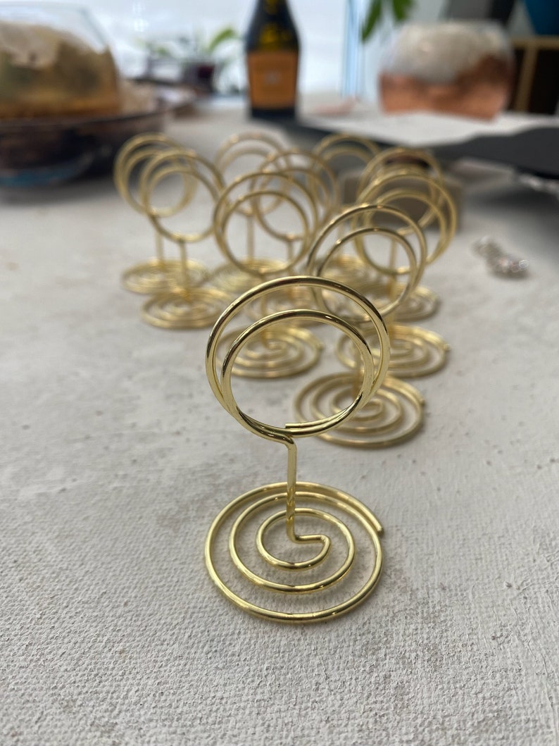 May include: A collection of gold-colored spiral place card holders. Each holder features a circular base with a spiral design and a wire loop for holding cards. The holders are arranged on a light-colored surface.
