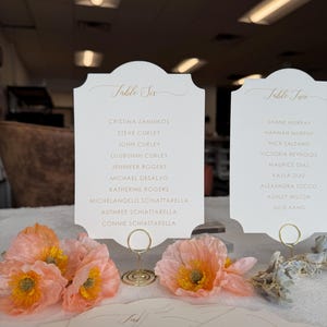 May include: Two white table cards with gold lettering. The card on the left is labeled "Table Six" and lists the names of guests. The card on the right is labeled "Table Two" and lists the names of guests.