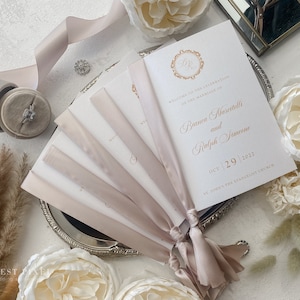 May include: A stack of wedding ceremony programs with a blush pink ribbon tied around them. The programs are on a silver tray with white flowers and dried grass. The front of the program features the text "Welcome to the celebration of the marriage of Bianca Marcitelli and Ralph Simeone Oct 29 2022 St. John's the Evangelist Church."