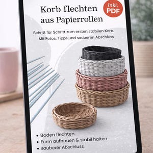 Op de afbeelding: Een tablet toont instructies voor het weven van manden van papieren rollen. De afbeelding toont een stapel geweven manden in zwart, grijs, roze en bruin, met een enkele mand op de voorgrond. De tekst op het scherm luidt "Korb flechten aus Papierrollen."