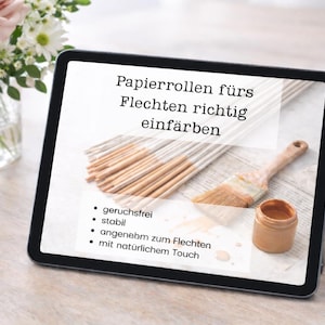 Op de afbeelding: Een tablet toont tekst in het Duits over het kleuren van papieren rollen. De afbeelding op de tablet toont penselen, verf en bundels houten stokken. Een vaas met roze rozen en witte bloemen, een mok en een notitieblok met een pen zijn ook zichtbaar.