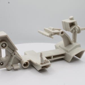 May include: A light gray plastic mechanical component with multiple articulated arms and joints. The object appears to be a part of a larger machine or device, with a complex, interlocking design. The component is set against a white background.