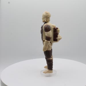 May include: A beige and brown action figure with a backpack. The figure has a light brown head and a dark brown body. The backpack is a dark brown color and has a light brown strap.
