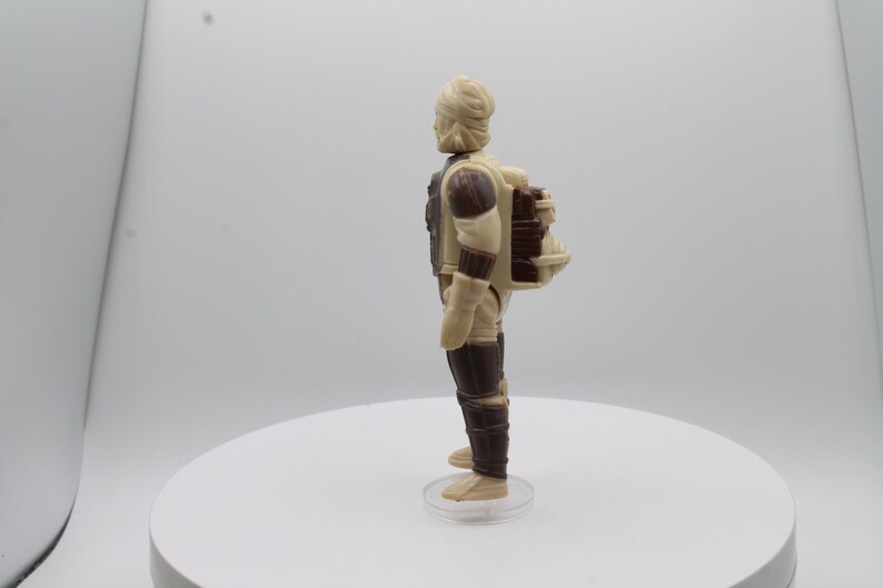 May include: A beige and brown action figure with a backpack. The figure has a light brown head and a dark brown body. The backpack is a dark brown color and has a light brown strap.