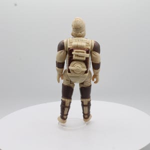 May include: A beige and brown action figure of a Star Wars character wearing a backpack and carrying a weapon. The figure is standing on a white surface.