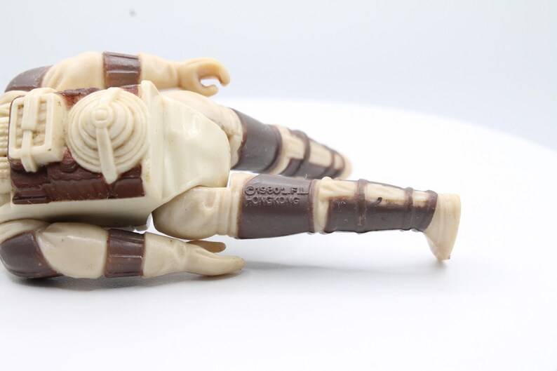 May include: A vintage Star Wars action figure of a Sandtrooper, lying on its side. The figure is made of plastic and has a brown and white color scheme. The figure has a backpack and a blaster rifle. The figure is marked with the copyright "&copy;1980 LFL" and "&copy;1980 Kenner".