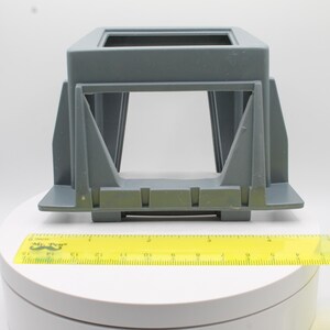 May include: A gray plastic rectangular object with an open center, possibly a support or housing. A yellow ruler with black markings is placed below the object, showing measurements in inches. The object has a geometric design.
