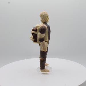 May include: A beige and brown action figure of a Star Wars character wearing a white head wrap and carrying a backpack.