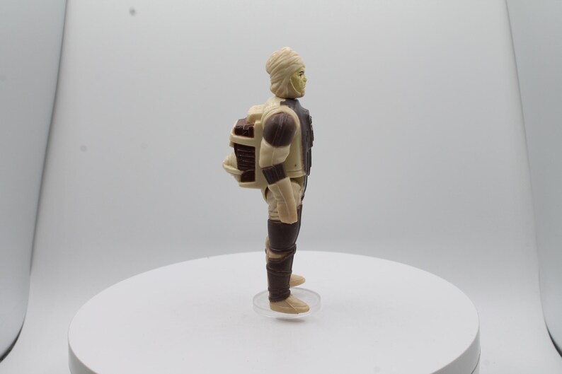 May include: A beige and brown action figure of a Star Wars character wearing a white head wrap and carrying a backpack.