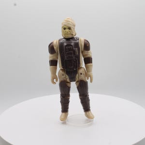May include: A vintage Star Wars action figure of a Sandtrooper with a brown and beige uniform and a white head wrap. The figure is standing on a white surface.