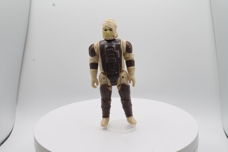 May include: A vintage Star Wars action figure of a Sandtrooper with a brown and beige uniform and a white head wrap. The figure is standing on a white surface.