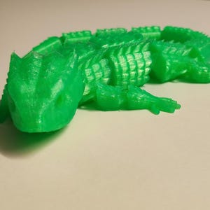 Könnte beinhalten: Eine grüne, 3D-gedruckte Eidechsenfigur mit einem strukturierten, segmentierten Körper. Die Eidechse ist in einer Pose mit dem Kopf zur Seite gedreht und leicht geöffnetem Maul.