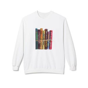 Könnte beinhalten: Weißes Sweatshirt mit einer Grafik von einem Stapel bunter Bücher, die mit Bindfaden zusammengebunden sind. Die Buchrücken zeigen Titel und Autorennamen, darunter "ABBY JIMENEZ OURS TRULY" und "The Friend Zone". Ein gemütliches, literarisches Design.