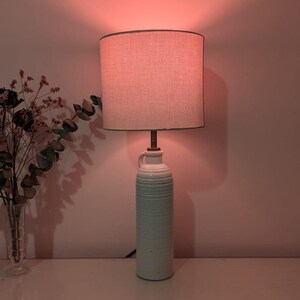Puede incluir: Una lámpara de mesa de cerámica con una pantalla de lino beige claro. La base de la lámpara es de color azul verdoso claro con un diseño texturizado y acanalado. La lámpara está sobre una superficie blanca, con un jarrón de flores secas a la izquierda.