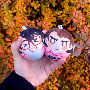 Puede incluir: Dos llaveros de peluche con caras de dibujos animados. Uno tiene gafas y una expresión sonriente, el otro una mirada severa. Los llaveros son rosas y blancos, con llaveros de metal plateado. El fondo está borroso con follaje otoñal.