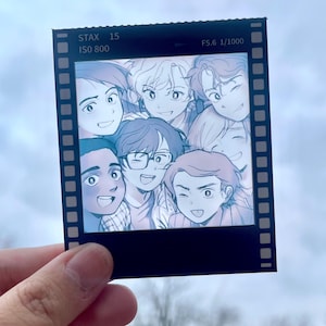 Könnte beinhalten: Ein schwarzes Foto in Form eines Filmstreifens mit einer Gruppe lächelnder Anime-Charaktere. Auf dem Foto steht oben "STAX 15 ISO 800 F5.6 1/1000". Das Bild wird von einer Hand vor einem verschwommenen Hintergrund im Freien gehalten.