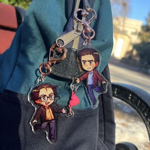 May include: Two clear acrylic keyrings featuring cartoon characters, attached to a rose gold-coloured clasp. One keyring depicts a character in a brown suit and glasses, the other in a maroon jacket. Both keyrings are hanging on a teal backpack.