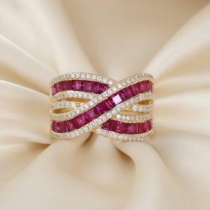 May include: A gold ring with a crisscross design, featuring rows of square-cut, deep red gemstones and small, sparkling white stones. The ring is set against a cream-colored, silky background.