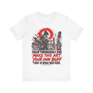 Peut inclure: Un t-shirt blanc avec une illustration graphique d'un samouraï tenant un sabre, un soleil rouge et le texte : TRAIN THOROUGHLY AND MAKE THIS ART YOUR OWN BODY THAT IS WHAT MATTERS. MIYAMOTO MUSASHI est inscrit en plus petit sous la citation.