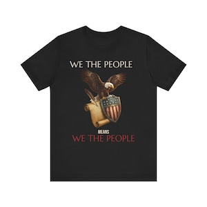Peut inclure: Un t-shirt noir à manches courtes avec un motif représentant un pygargue à tête blanche, une plume, un parchemin et un bouclier aux couleurs du drapeau américain. Le texte WE THE PEOPLE et MEANS WE THE PEOPLE est imprimé en blanc et rouge.