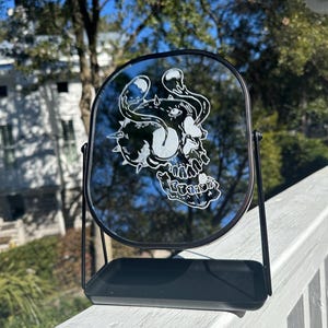 Może przedstawiać: Czarne lustro stołowe z okrągłym, fazowanym szkłem. Lustro ma wygrawerowany biały wzór czaszki z rogami i spiralnym wzorem. Lustro stoi na czarnym stojaku z małą półką.