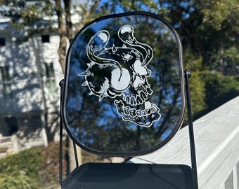 以下が含まれることがあります： 丸い面取りされたガラス面を持つ黒い卓上ミラー。ミラーには、角と渦巻き模様のある頭蓋骨の白くエッチングされたデザインが施されています。ミラーは、小さな棚が付いた黒いスタンドの上に置かれています。