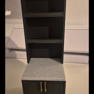 Op de afbeelding: Een zwarte miniatuurkeukenkast met een grijs aanrechtblad en twee deuren. Boven het aanrechtblad bevinden zich twee planken. De tekst "Larder Option" staat op de voorkant van de kast.