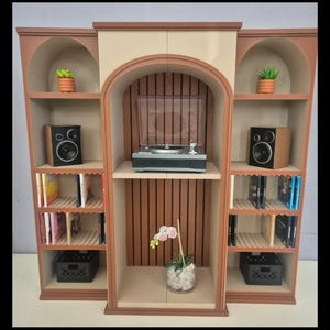 May include: A miniature three-section shelving unit with arched tops, featuring a central record player, two speakers, small potted plants, books, and black storage bins. The structure is finished in light beige and brown tones with vertical slats.
