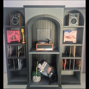 Op de afbeelding: Een grijze houten media-opbergeenheid met drie boogvormige secties. De zijplanken bevatten luidsprekers, een gouden microfoontrofee en vinylplaten. Het middengedeelte is voorzien van een houten platenspeler en een kleine plant in een pot.
