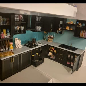 Puede incluir: Miniatura de cocina con gabinetes oscuros, un salpicadero turquesa y una encimera blanca. El conjunto incluye una estufa, un fregadero y varios accesorios de cocina, como alimentos, utensilios y electrodomésticos. Los gabinetes tienen manijas doradas.