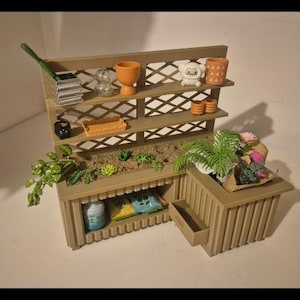 Può includere: Un'esposizione di giardino in miniatura con una struttura in legno marrone con ripiani, un'aiuola e un piccolo cassetto. I ripiani contengono vasi in miniatura, un annaffiatoio e oggetti decorativi.