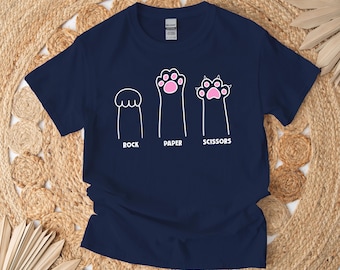 Può includere: Una maglietta blu navy con una grafica bianca di tre zampe di gatto che rappresentano sasso, carta, forbice. Le zampe hanno i cuscinetti rosa.